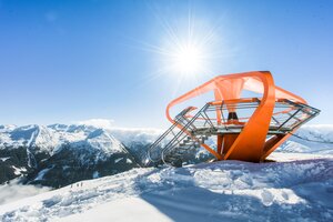 An orange viewing platform is surrounded by snow and a snow-covered mountain panorama | © Gasteiner Bergbahnen AG, Wolkersdorfer