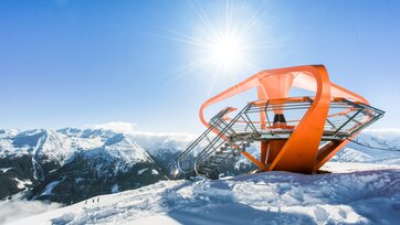An orange viewing platform is surrounded by snow and a snow-covered mountain panorama | © Gasteiner Bergbahnen AG, Wolkersdorfer