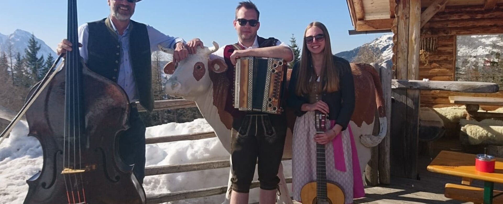 Drei Musiker in Tracht mit Instrumenten vor Almhütte und Holzkuh in verschneiter Kulisse