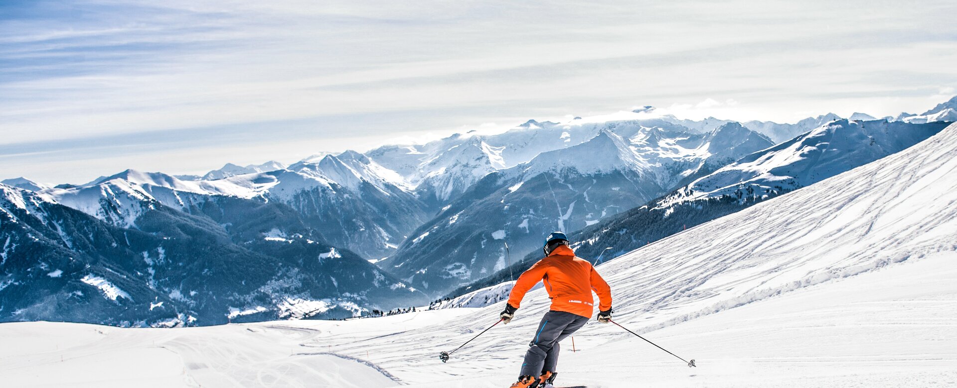 Skier with orange ski jacket descends a groomed slope and snow-capped mountains can be seen in the distance | © Gasteinertal Tourismus GmbH, Creatina