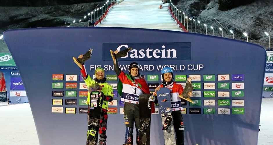 Three snowboarders celebrate on the World Cup podium in Gastein, slope lit up in the background. | © KTVB Bad Gastein, Simon Hutter