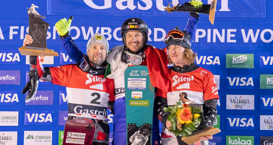 Three snowboarders celebrate on Gastein podium, holding trophies, flowers, and their boards. | © KTVB Bad Gastein, Magotree Photography
