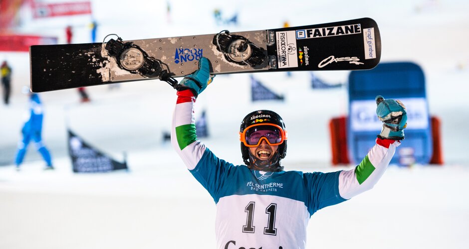 Snowboarder with bib number 11 celebrates with arms raised, holding board overhead in Gastein. | © FIS, Miha Matavz