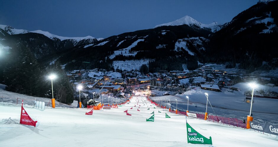 Lit-up racecourse with slalom gates descends into Bad Gastein beneath evening mountain scenery. | © KTVB Bad Gastein, Simon Hutter