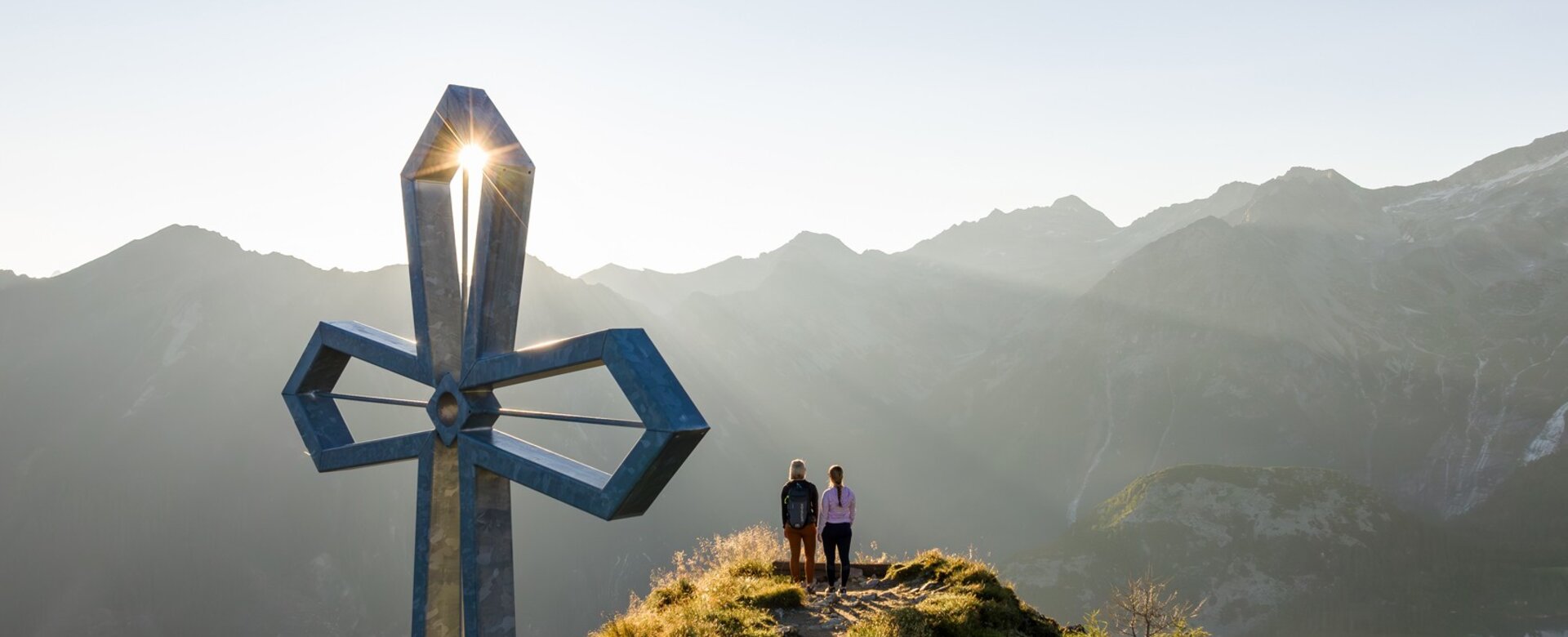 Two hikers at summit cross during sunrise with alpine panorama in the background