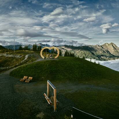 Heart-shaped wooden swing on a hill, mountains and sea of clouds in the valley, dramatic clouds in the sky. | © Großarler Bergbahnen