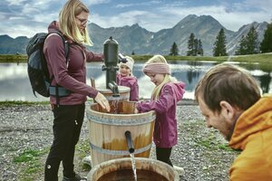 Family with kids pumping water from wooden barrels by a lake in Großarl, mountains and clouds reflecting in the background. | © Großarler Bergbahnen