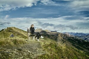 Woman sits on a bench along a mountain path in Großarl, overlooking autumn hills and a dramatic cloudy sky. | © Großarler Bergbahnen
