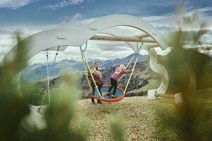Two kids play on a large nest swing with a mountain backdrop in Großarl, wood chips on the ground, clouds in the sky. | © Großarler Bergbahnen