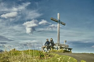 Two people rest on a bench by the summit cross in Großarl, enjoying the view beneath a dramatic, partly cloudy sky. | © Großarler Bergbahnen