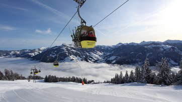 Yellow gondolas with skiers ride over snowy slopes, sea of clouds below, mountains in the background, clear sky.