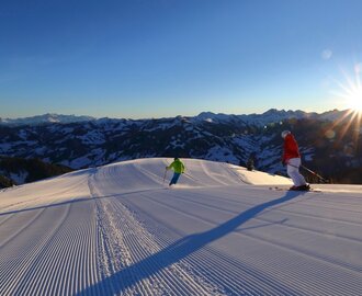 Zwei Skifahrer auf frisch präparierter Piste bei Sonnenaufgang mit Alpenpanorama