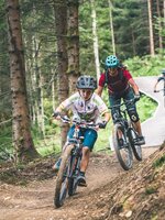 Mountain bikers ride narrow flow trail through forest with wooden bridges and bends