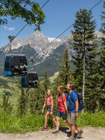 Family hiking on forest trail with gondolas in background and mountain panorama | © Roland Haschka