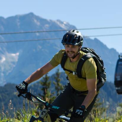 A mountain biker rides a winding trail with a gondola lift and scenic alpine peaks in the background under a clear blue sky. | © Roland Haschka