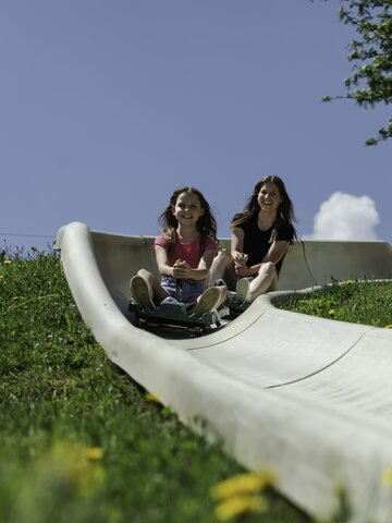 Girl and woman ride down the Biberg summer toboggan track, smiling in the sun, with flowers and blue sky all around. | © Hochkönig / Roland Haschka