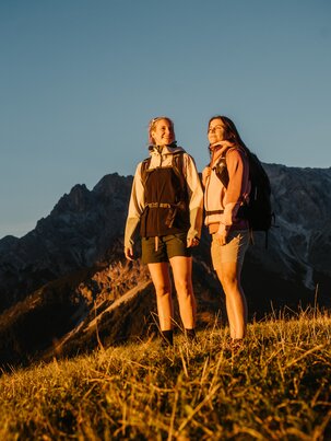 Zwei Wanderinnen stehen im Abendlicht auf einer Wiese in Dienten vor einem felsigen Bergmassiv nahe der Gabühelbahn. | © Hochkönig Bergbahnen