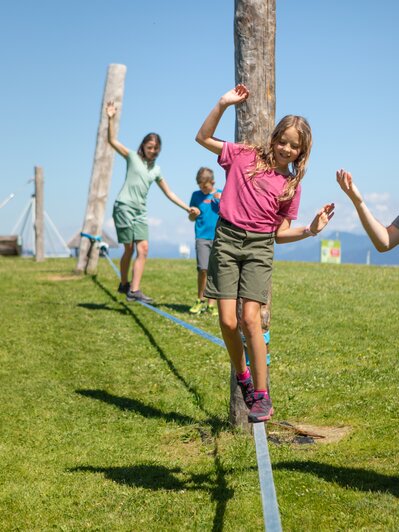 A girl balances on a slackline in Mühlbach while a man helps and two children follow behind. | © Hochkönig Bergbahnen, Roland Haschka