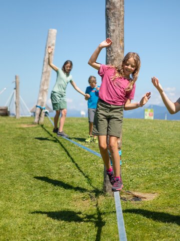 A girl balances on a slackline in Mühlbach while a man helps and two children follow behind. | © Hochkönig Bergbahnen, Roland Haschka