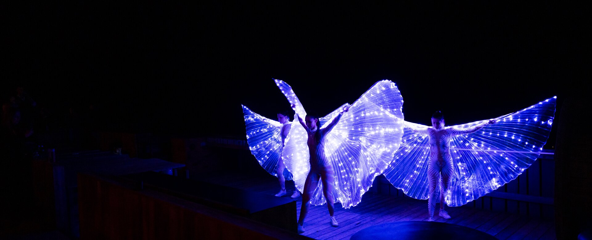 Three performers with LED wing costumes glowing in the dark on wooden terrace of mountain hut