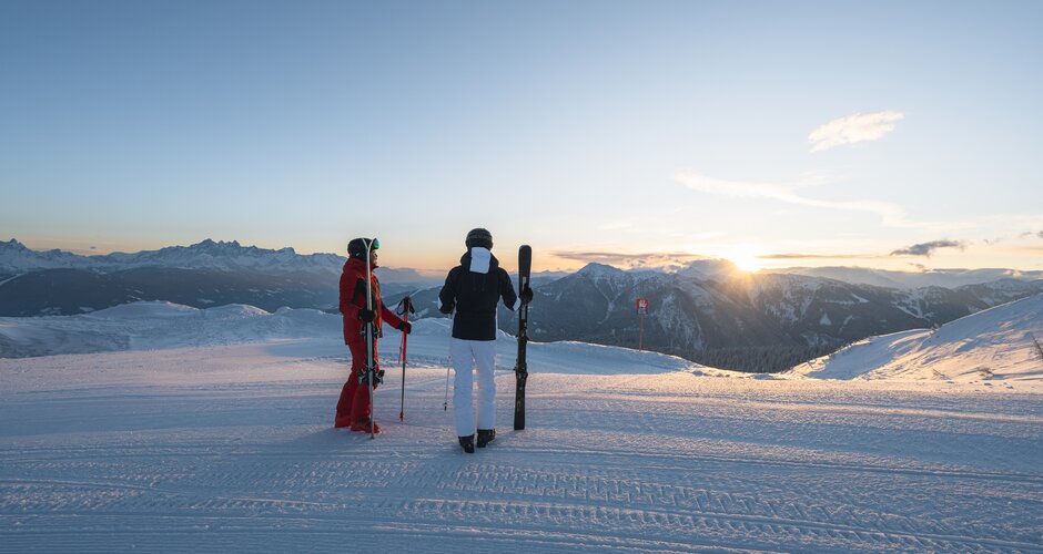 A person in a red ski suit and a person in a white and black one are standing on a slope watching the sunrise | © Ski amadé