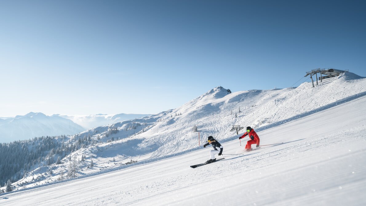Two skiers ski down the freshly prepared piste and in the background you can see the top station of a chairlift | © Ski amadé