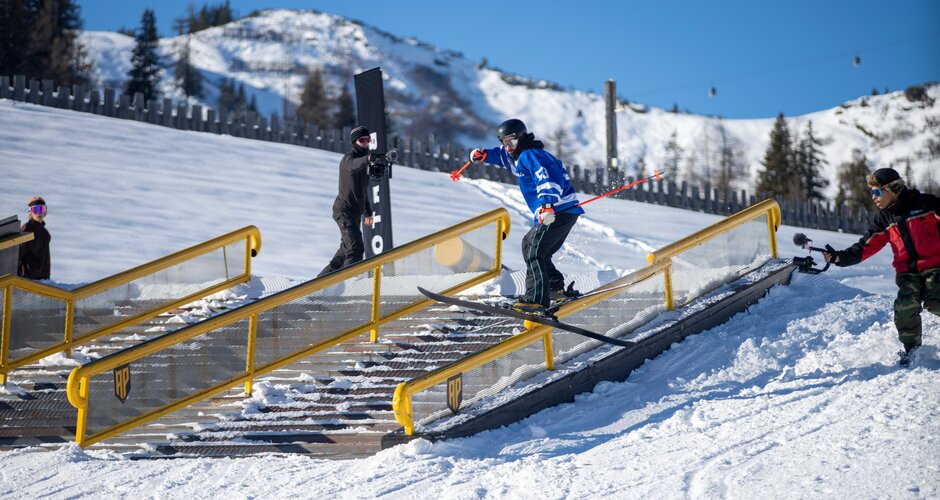Snowboarder jumps onto a yellow railing while a cameraman stands on the right side filming the snowboarder | © Markus Fischer