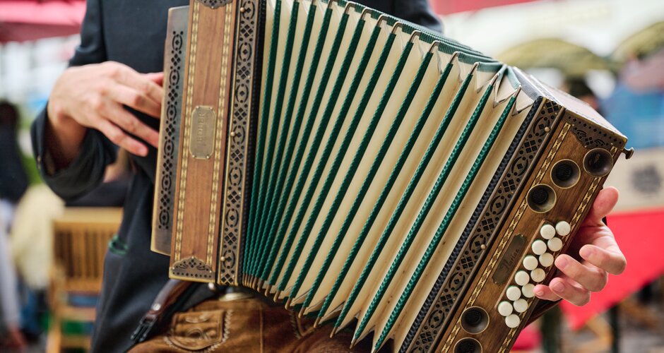 A light brown-green accordion is played by a seated person in lederhosen | © Matthias Fritzenwallner