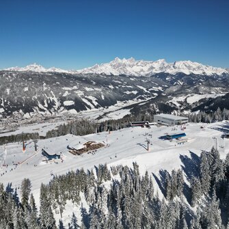 A gondola station, two chairlift stations and a ski hut from above and many houses can be seen in the valley below