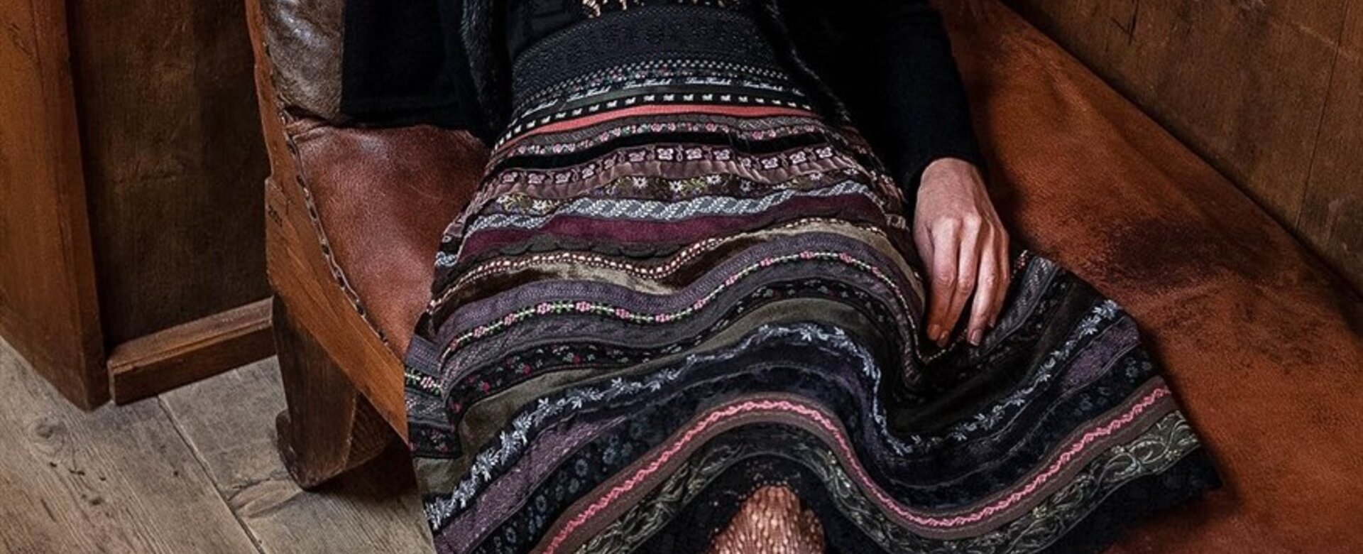 Frau in traditionellem Rock mit Muster sitzt auf braunem Ledersofa in Holzzimmer.