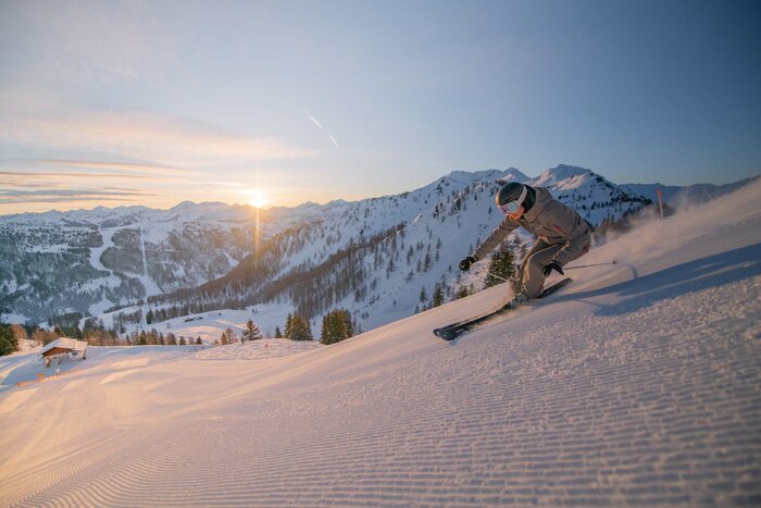 Skier in a beige ski suit skiing down a freshly groomed piste as the sun rises behind the mountains.