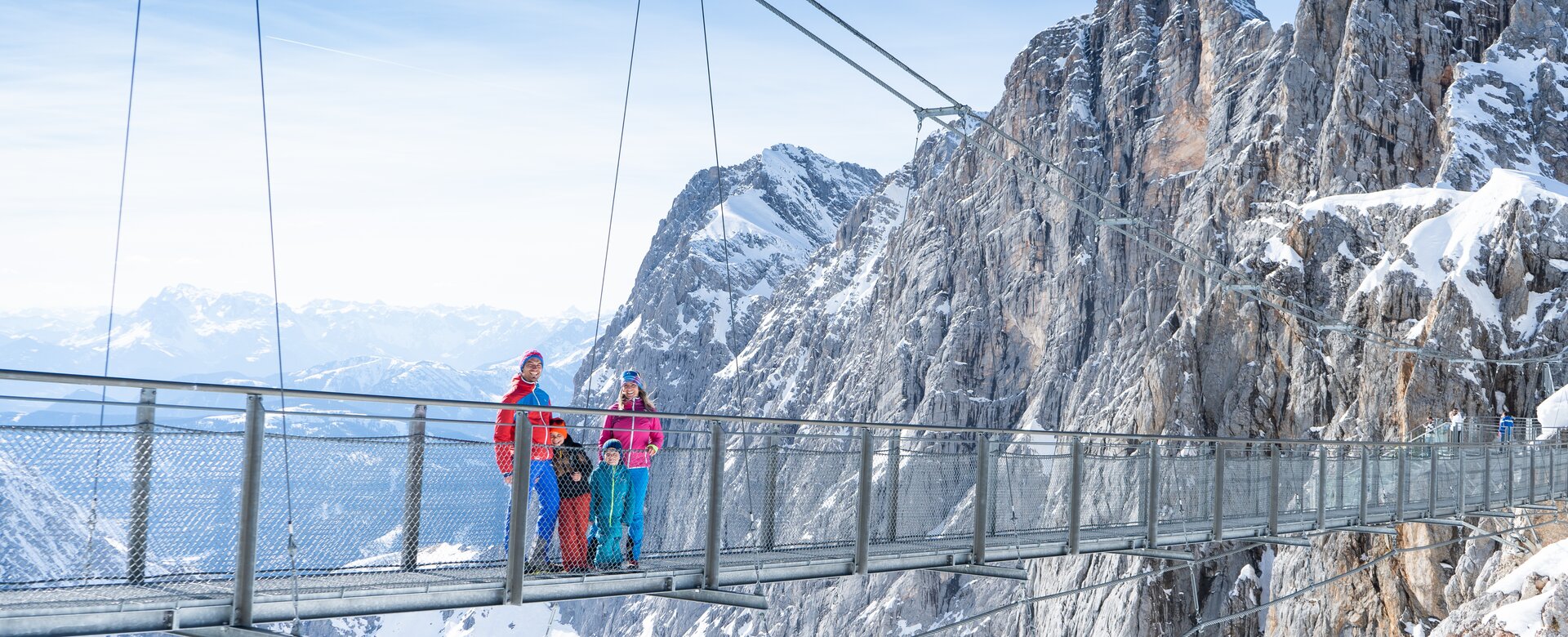 Zwei Erwachsene und zwei Kinder stehen auf einer Hängebrücke und unten sieht man verschneite Berge | © Josh Absenger
