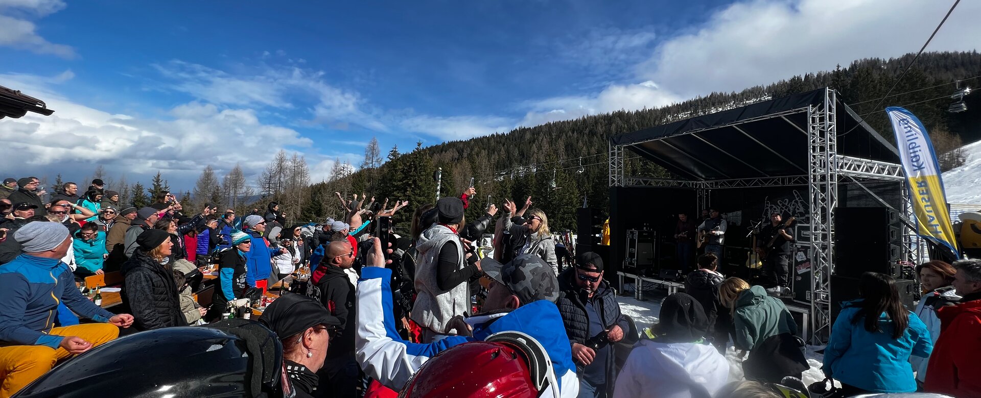 Many people in ski gear stand in front of a small stage on which musicians play. | © Hauser Kaibling