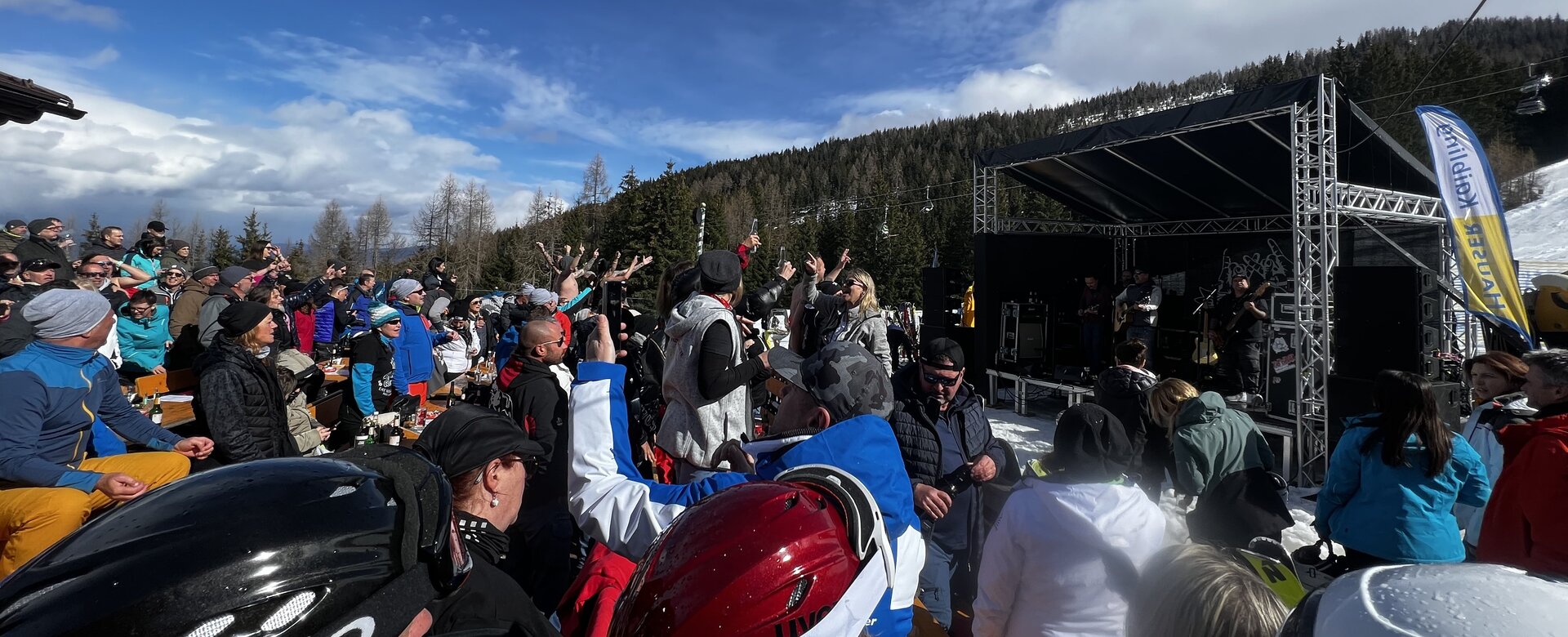 Many people in ski gear stand in front of a small stage on which musicians play. | © Hauser Kaibling