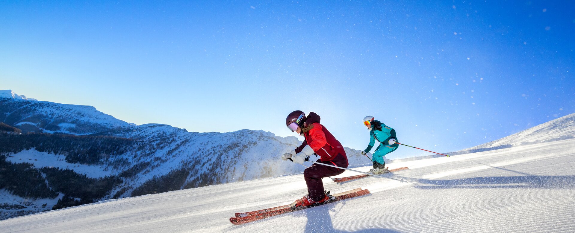 Two skiers, one in blue and the other in red, ski down the groomed piste with mountain peaks in the background