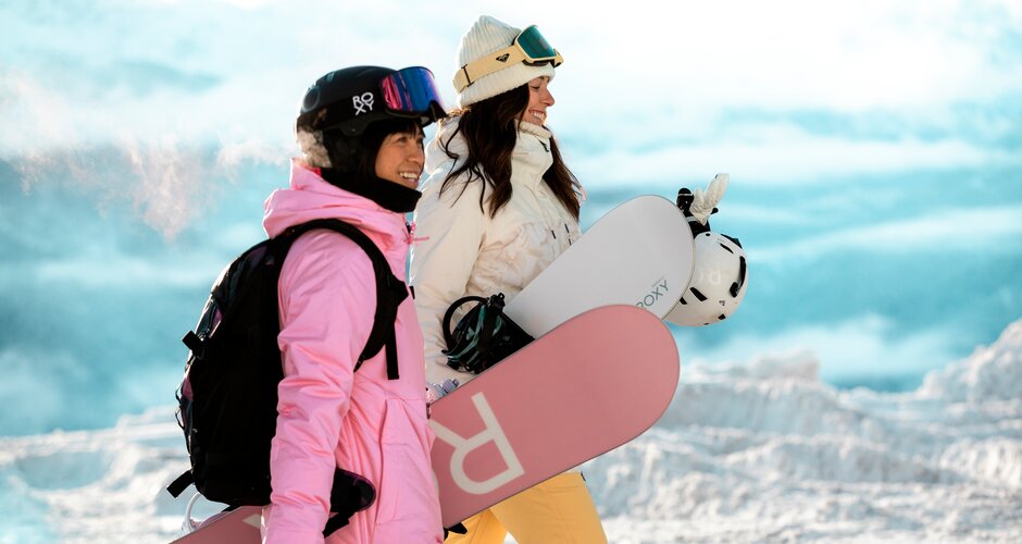 Two young ladies with snowboard equipment walk past the photographer on the piste and to the side | © QParks