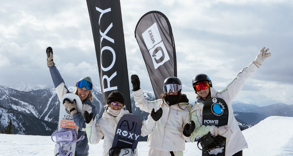 Four female snowboarders pose in snow with boards and flags, snowy mountains in the background | © Henrie Kelbing