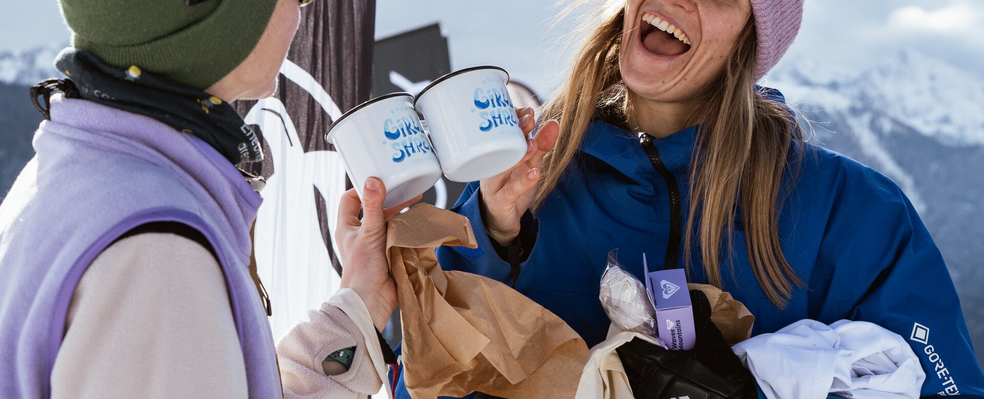 Two women toast with white enamel mugs, laughing in front of snowy mountain scenery. | © Henrie Kelbling