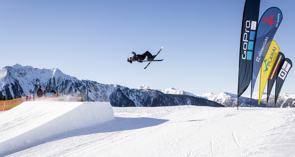 Freestyle skier jumps over snow kicker at Superpark Planai with flags and mountains in background | © Hannes Mautner
