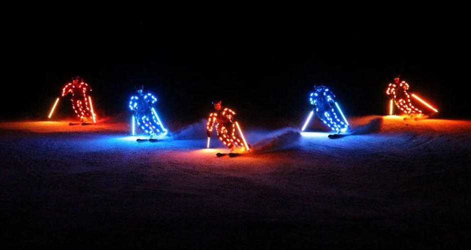 5 skiers with light elements on their ski suits and ski poles ski down the piste in a formation | © Erlebnis Rittisberg
