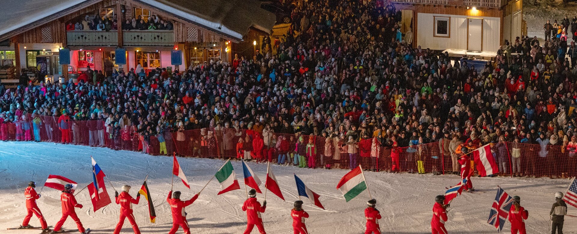 Skiers in red carry national flags on a slope at night in front of a large crowd | © Michael Simonlehner