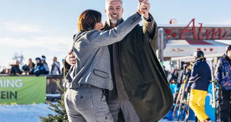 A man and woman dance in winter fashion in front of the Almbar on snowy Reiteralm mountain | © Gerald Grünwald