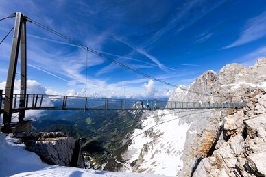 People cross a suspension bridge on snowy Dachstein with a wide view over green valley under a clear blue sky. | © Harald Steiner
