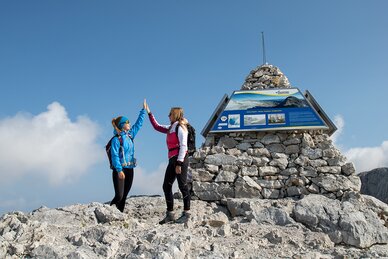 Zwei Frauen in Wanderkleidung geben sich auf einem felsigen Gipfel ein High-Five neben einer Infotafel auf einem Steinhaufen. | © Birgit Walcher