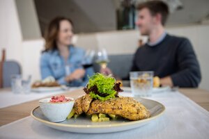 Nahaufnahme eines panierten Fischfilets mit Salat und Beilage, im Hintergrund unscharf lachendes Paar mit Weingläsern. | © Harald Steiner