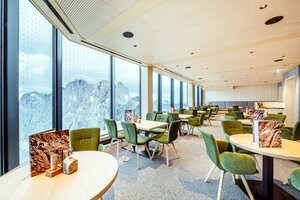 Bright restaurant with large panoramic windows, green upholstered chairs, and wooden tables overlooking the surrounding mountains. | © Christoph Huber