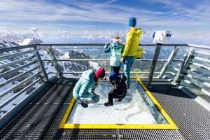 Familie steht auf dem Sky Walk mit Glasboden am Dachstein und blickt auf verschneite Berge und grüne Täler. | © Harald Steiner