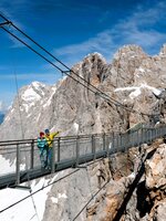 Two people cross the Dachstein suspension bridge with snowy peaks and rugged cliffs in the background under clear blue skies. | © Harald Steiner
