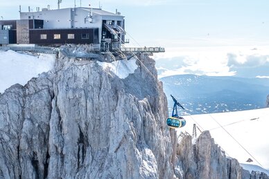 Eine blaue Seilbahn fährt zur modernen Bergstation auf schroffen Felsen, mit weitem Blick auf schneebedeckte Gipfel und Täler. | © Josh Absenger