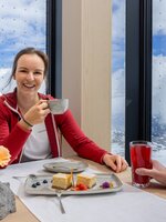 Smiling woman in a red jacket drinks coffee, man with red drink, Alpine panorama through large windows, dessert on table. | © Harald Steiner